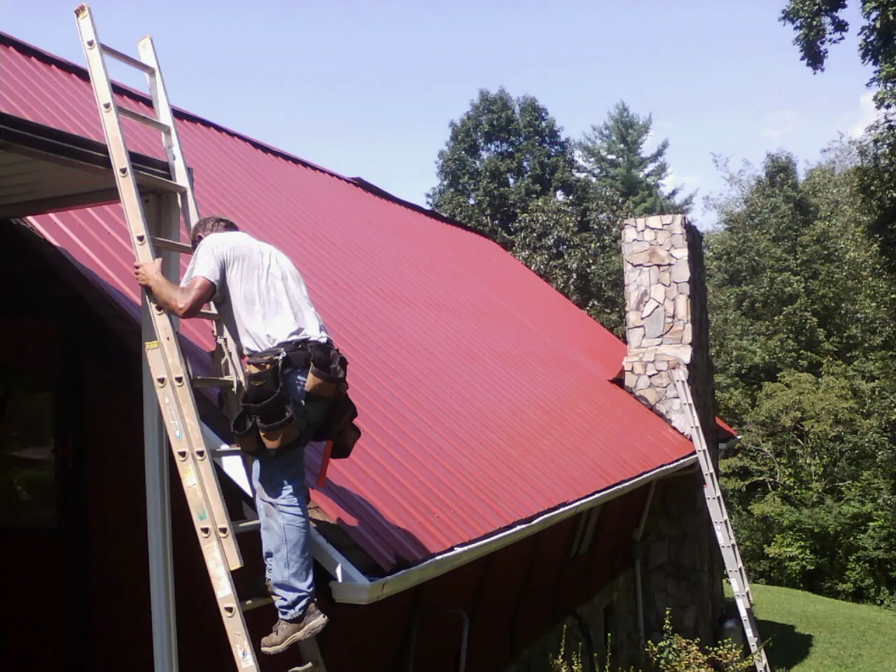 Roofer on a ladder beside a red metal roof and stone chimney — Soffit & Fascia Repair by Roofing Brothers in Redkey